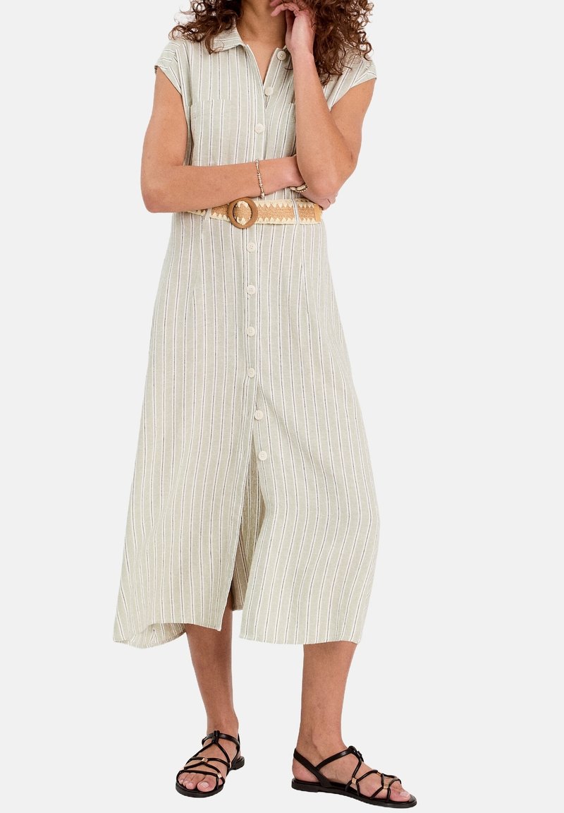 Femme portant une robe midi beige à rayures avec boutons, une ceinture tissée et des sandales plates noires à brides, debout les bras croisés.