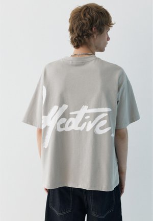 Jeune homme aux cheveux mi-longs portant un t-shirt beige ample avec un grand texte blanc "Native" dans le dos et un pantalon foncé.