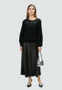 Schwarzer gestrickter Pullover mit einem Perlenmuster am Ausschnitt, kombiniert mit einem schwarzen Satinkleid, schwarzen High Heels und einer silbernen Handtasche.