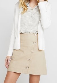 Gonna beige con chiusura a bottoni e tasca laterale, abbinata a un cardigan bianco a coste su una camicia a quadri grigia chiara.