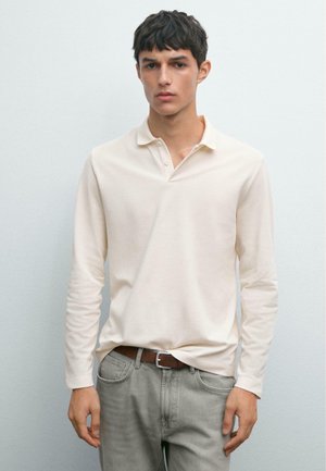 Young man wearing a cream long-sleeve polo shirt tucked into light gray jeans with a brown belt, standing against a plain light wall.