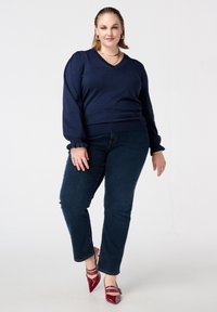 Marineblauwe gebreide trui met een V-hals en ballonmouwen, gecombineerd met donkere denim jeans en rood lakleren pumps met een spitse neus.