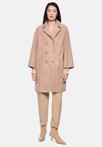 Doppelreihiger beiger Wildledermantel mit breitem Kragen, Seitentaschen und lockeren Ärmeln, kombiniert mit hellbeigen Hosen und passenden spitzen Absätzen.