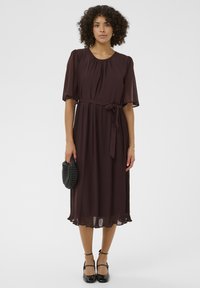 Ciemnobrązowa sukienka z plisami, krótkimi bufkami, dopasowanym paskiem w talii oraz falbaniastym dołem, w zestawie z teksturowanym czarnym kopertówką i czarnymi szpilkami.