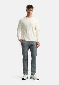 Helle beige Langarmbluse aus strukturiertem Stoff, kombiniert mit grauen gemusterten Hosen und weißen Sneakers mit grauen Akzenten.