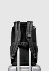 Mochila negra de cuero con correas acolchadas de malla, bolsillos laterales y una hebilla de seguridad, descansando sobre una maleta plateada contra un fondo claro.