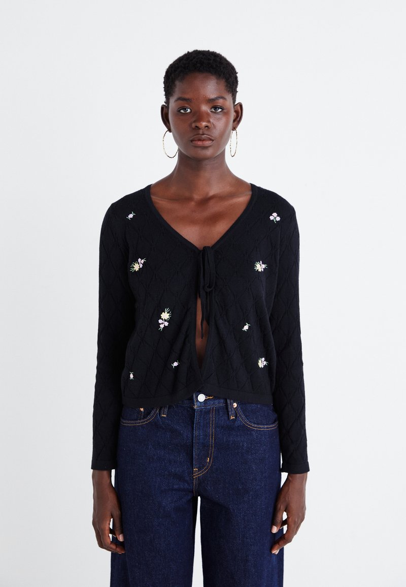 Cardigan negru tricotat cu model de diamant, cu broderii florale și șnur în față. Mâneci lungi, croială scurtă și decolteu deschis.