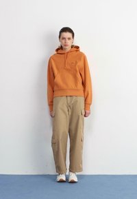 Orange hooded sweatshirt with a front pocket, paired with beige cargo pants featuring side pockets. Worn with white and tan sneakers.