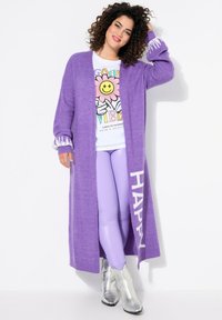Długi fioletowy kardigan z białymi akcentami na mankietach i napisem "HAPPY", zestawiony z białym t-shirtem z grafiką oraz błyszczącymi lawendowymi legginsami, zakończony srebrnymi botkami.
