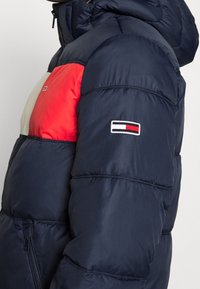 Granatowa kurtka puchowa z czerwoną i kremową panelem na klatce piersiowej, z materiałową łatą z logo na rękawie oraz kieszenią na zamek błyskawiczny z przodu.