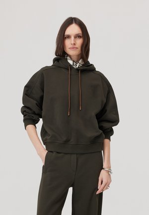 Kobieta w ciemnozielonej, oversize’owej bluzie z kapturem i pasujących spodniach, stojąca z jedną ręką w kieszeni na jednolitym jasnym tle.