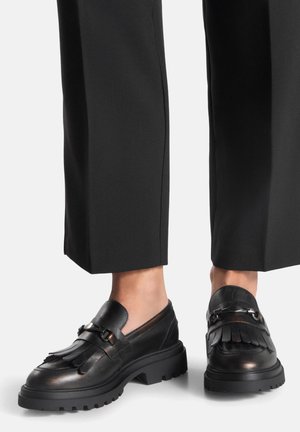 Schwarze Leder-Loafers mit Fransen, dicken Gummisohlen und dekorativem Beschlag, kombiniert mit maßgeschneiderten schwarzen Hosen.