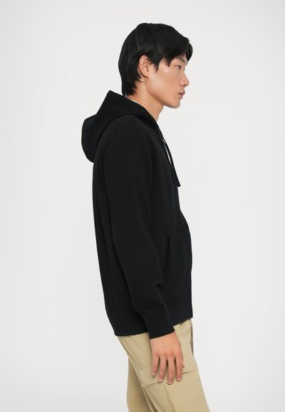 Sweatshirt preto com capuz, bolso frontal e cordão. Feito de tecido macio, apresenta um corte descontraído e punhos canelados, mostrado com calças caqui.