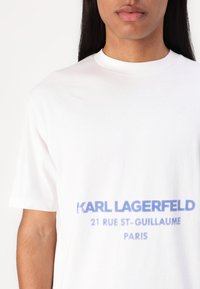 Λευκή βαμβακερή μπλούζα με χαρακτηριστικό μπλε κείμενο: "KARL LAGERFELD 21 RUE ST-GUILLAUME PARIS" στο μπροστινό μέρος, στρογγυλή λαιμόκοψη, κοντά μανίκια.