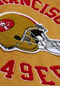 Tessuto giallo con logo ricamato dei San Francisco 49ers. Presenta un elmo da football dorato con dettagli bianchi e neri e scritte rosse.