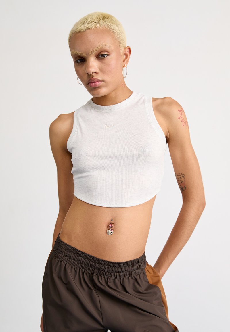 White ribbed crop top with a high neckline and Nike logo. Model wears brown drawstring trousers with an elastic waistband.