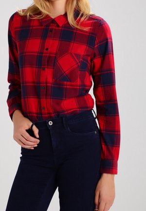 Femme portant une chemise à carreaux rouge et marine boutonnée, rentrée dans un jean taille haute bleu foncé, la main reposant près de la ceinture.