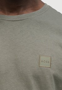 Λαδί πράσινο βαμβακερό μπλουζάκι με στρογγυλή λαιμόκοψη και μια μικρή, τετράγωνη, ραμμένη ετικέτα με το γράμμα "BOSS" σε λευκό χρώμα.