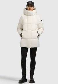 Fehér puffertöltetű kabát magas gallérral és steppelt mintával, fekete leggings-szel és katonai csizmával párosítva. Fekete sapka logó részlettel.