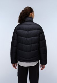 Svart puffjakke med chevron-søm, høy krage og teksturert finish. Bært over en stripete skjorte og svarte bukser, som gir et avslappet utseende.