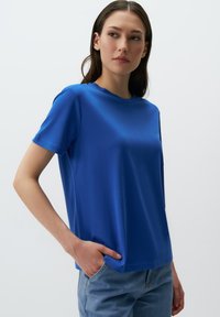 Niebieska koszulka polo z krótkim rękawem z miękkiego materiału, z okrągłym dekoltem, luźnym krojem i detalami stębnowania na rękawach oraz u dołu.