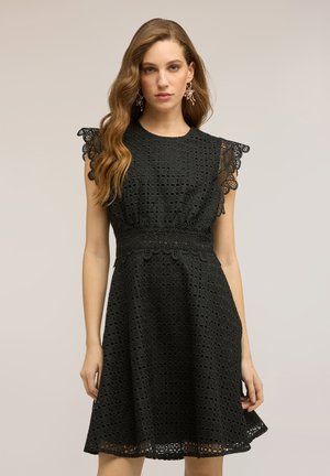 Frau mit langem welligem Haar trägt schwarzes ärmelloses Spitzenkleid mit wellenförmigen Kanten, steht vor einem schlichten neutralen Hintergrund.