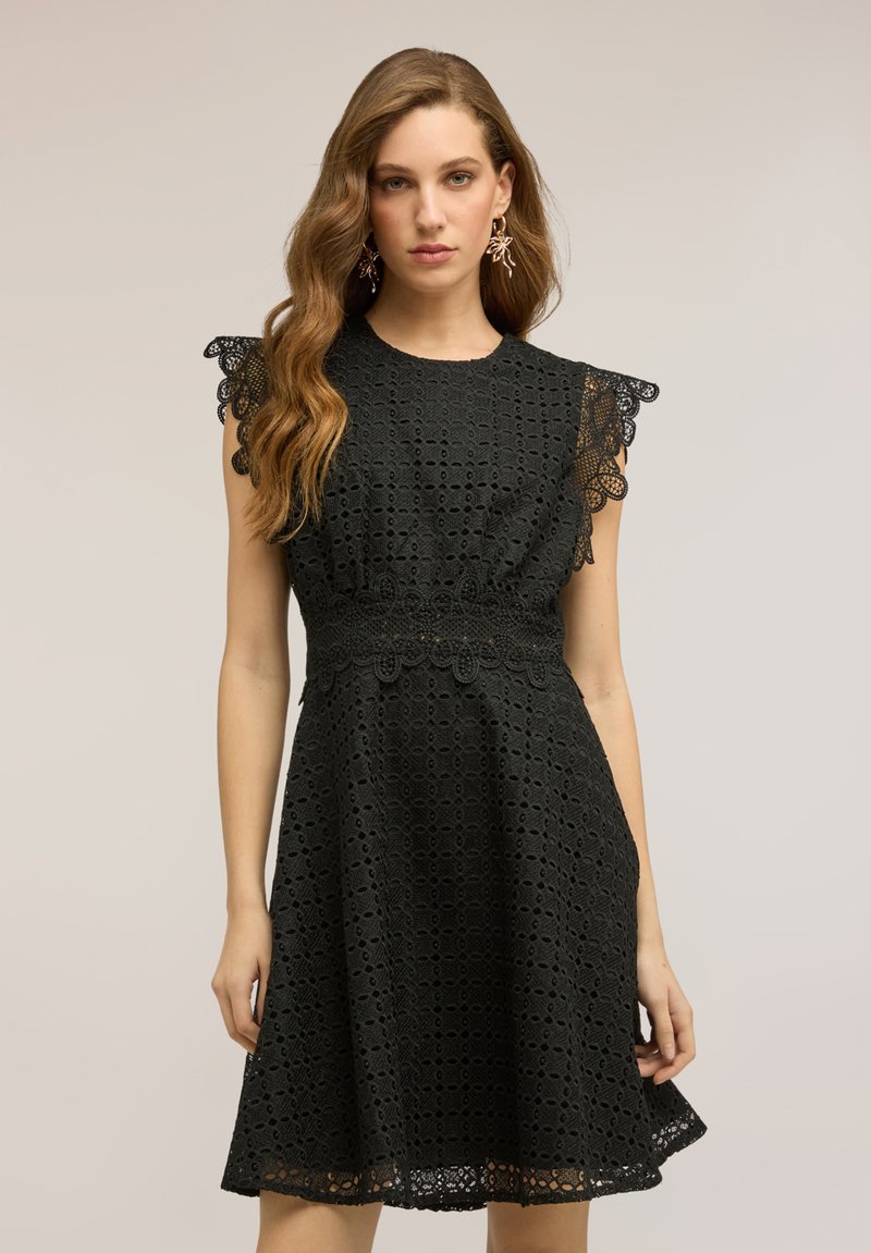 Femeie cu păr lung și ondulat poartă o rochie neagră fără mâneci din dantelă cu margini crestate, stând în fața unui fundal simplu și neutru.