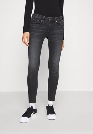 Persoon in zwarte skinny jeans, gecombineerd met zwart-witte sneakers en witte sokken, staand tegen een effen achtergrond.