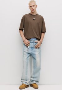Hnědé oversized tričko, světle modré volné džíny se sepráním, tanové šněrovací boty. Doplňky zahrnují prsteny, náušnice a dekorativní náhrdelník.