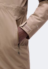 Main à la peau foncée insérée dans la poche zippée de la manche d’une veste imperméable beige avec une sangle Velcro réglable portant la mention DRYVENT.