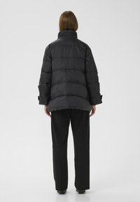 Sort puffjakke med høj krave, der har quiltede sektioner og en let oversized pasform. Bæres over sorte bukser.