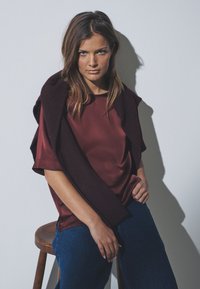 Bordeauxfarbenes Satinoberteil mit kurzen Ärmeln, drapiert mit einem dunklen Schal; kombiniert mit blauen Jeans mit weitem Bein. Modell sitzt auf einem hölzernen Hocker.