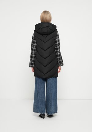 Femme portant un gilet matelassé noir à capuche sur une chemise à carreaux noire et blanche et un jean taille large bleu, se tenant contre un fond blanc uni.