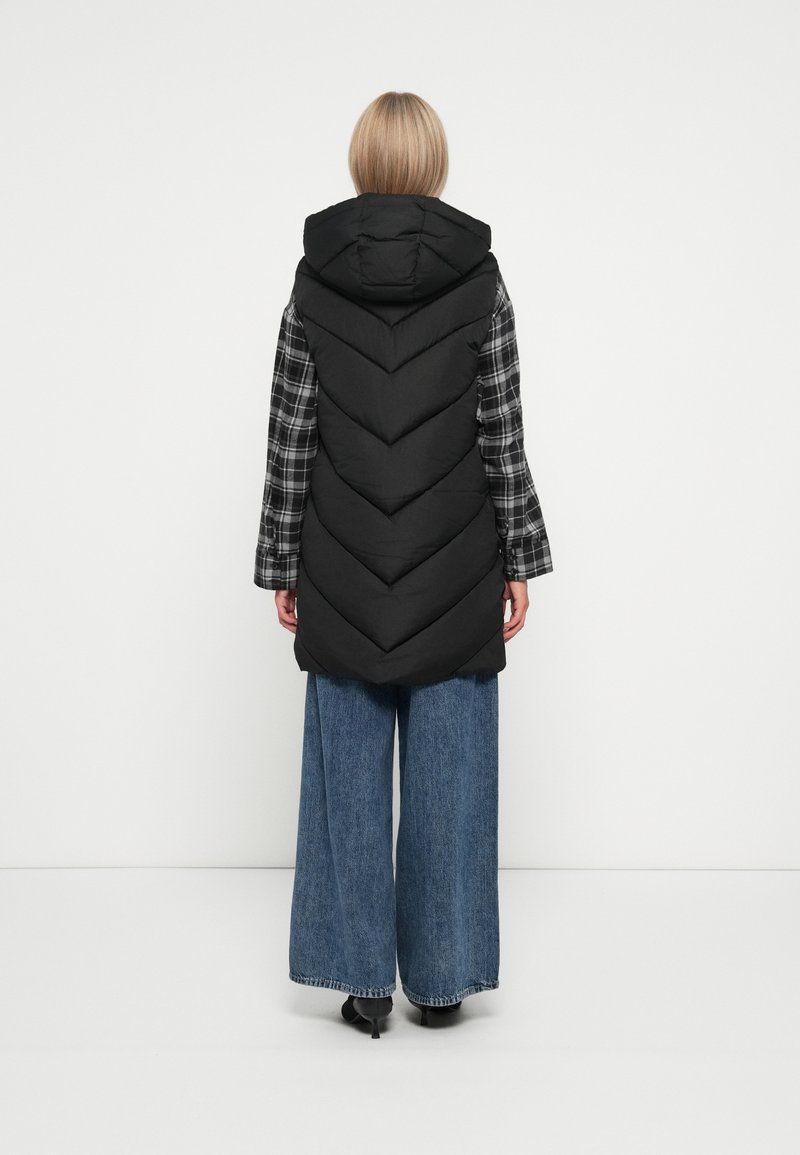 Kvinne iført en svart hettejakke over en svart og hvit rutete skjorte og vide blå jeans, stående mot en ensfarget hvit bakgrunn.