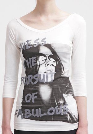 Mujer que lleva una camiseta blanca ajustada de manga larga con una imagen en blanco y negro de una persona con gafas de sol y el texto "Guess The Pursuit of Fabulous."