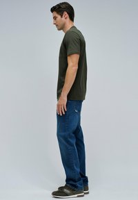 Hombre de pie de lado, vistiendo una camiseta verde oliva, jeans azules y zapatillas oscuras, frente a un fondo claro y sencillo.