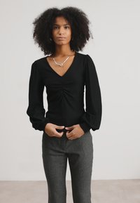Black long-sleeve top with a V-neck, featuring a gathered center design, paired with grey textured trousers.