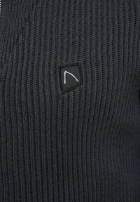 Schwarzer, gerippter Strickpullover mit einem Logopatch in weißem Design. Die Textur ist dick und gewoben, mit einer taillierten Silhouette.