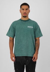 Groen oversized shirt met korte mouwen en een vervaagde textuur, ronde halslijn en een klein wit logo op de linkerborst. Zwarte broek die onder het shirt wordt gedragen.