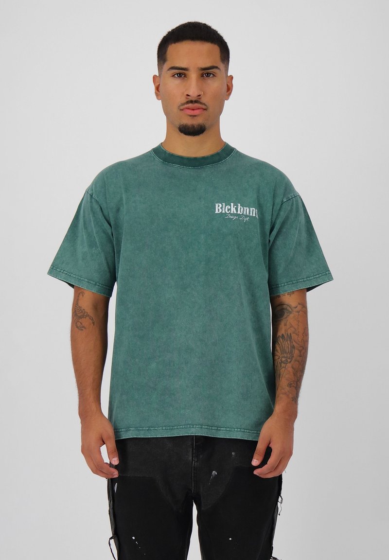 Groen oversized shirt met korte mouwen en een vervaagde textuur, ronde halslijn en een klein wit logo op de linkerborst. Zwarte broek die onder het shirt wordt gedragen.
