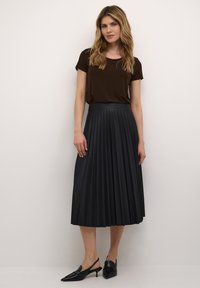Fekete, pliszírozott midi szoknya sima derékkal, amelyet egy rövid ujjú barna toppal párosítanak. A lábbeli fekete, hegyes orrú slingback magassarkú.