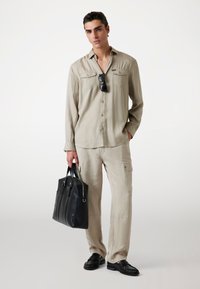 Beige overhemd met lange mouwen en twee borstzakken, gecombineerd met beige cargo-broeken. Donkere zonnebril en zwarte leren tote bag. Zwarte instappers.