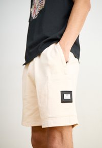 Light beige shorts made of cotton blend, featuring a side cargo pocket with a black label patch. Soft fabric with a smooth texture.