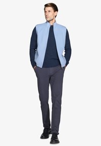 Gilet in pile azzurro chiaro da uomo con chiusura a zip sopra un maglione navy, abbinato a pantaloni grigio scuro e scarpe nere. Texture morbida, design casual.