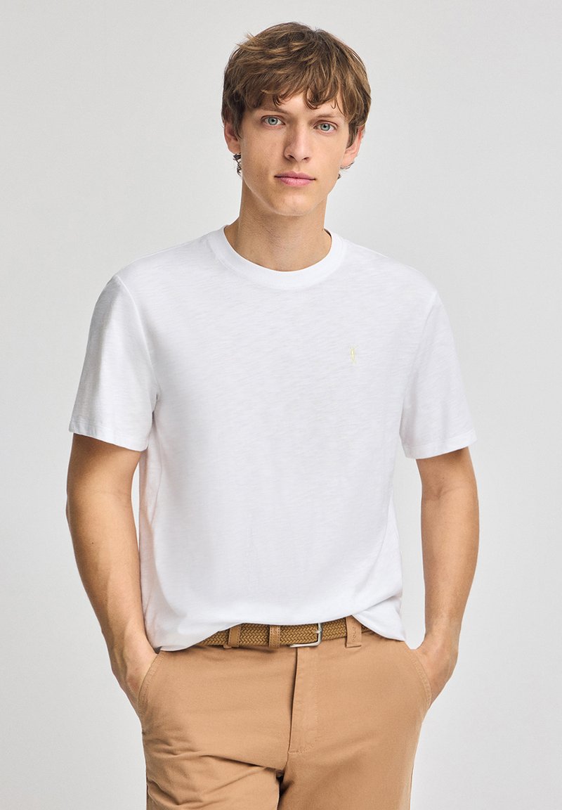 Vit kortärmad t-shirt med rund hals och liten gul logodetalj på bröstet, tillsammans med beige byxor och ett brunt bälte.