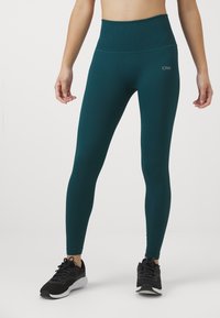 Hoge taille teal leggings met ribbelstructuur en subtiele logo aan de voorkant, gecombineerd met zwarte sportschoenen.