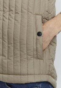 Gilet beige trapuntato con cuciture verticali, dotato di una tasca laterale chiusa da un bottone nero e un orlo arrotondato.