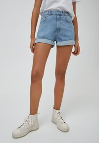 Person med uppvikta ljusblå denimshorts, vita högklackade sneakers och en vit t-shirt, som står mot en enkel ljus bakgrund.