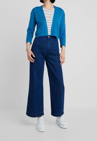 Blue cropped cardigan over a striped shirt, paired with high-waisted, wide-leg dark jeans and white sneakers. Simple design, casual fit.