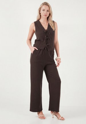 Blondína v čiernej bezrukávovej veste so zaväzovaním vpredu a zodpovedajúcich širokých nohaviciach, béžových sandáloch na opätku a vrstvených zlatých náhrdelníkoch.